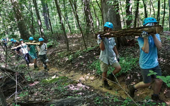 A group of students wearing gloves and helmets carry stumps along a trail in a wooded area. 